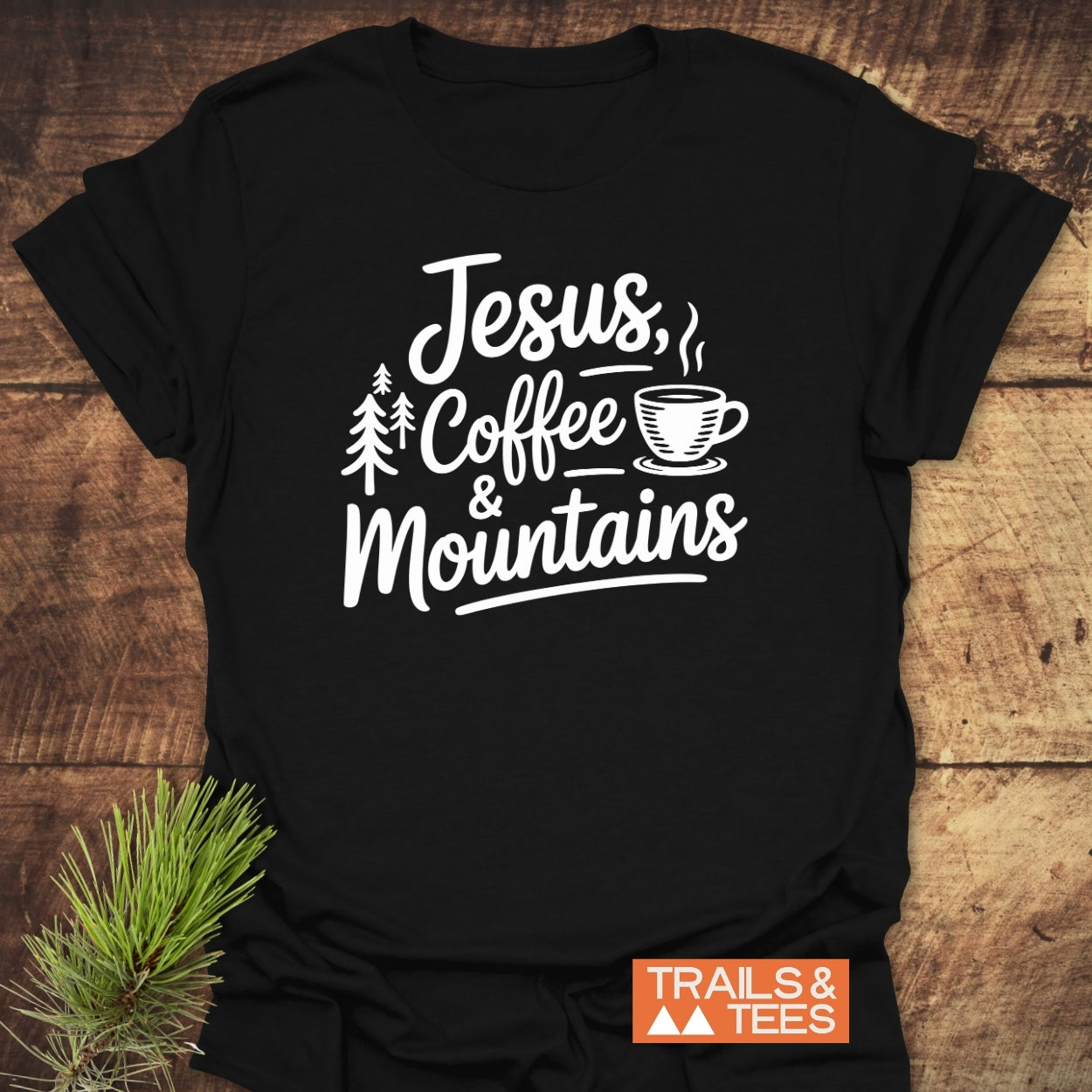 Jesus, Coffee, Mountains T-Shirt featuring white text and graphics of pine trees and a coffee cup on black fabric. Laid flat on a wooden surface, with “TRAILS & TEES.” logo in the corner.