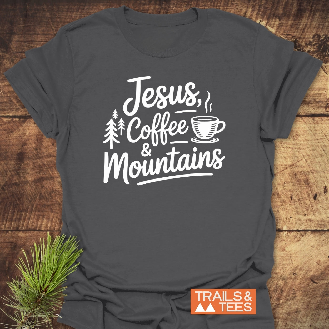 Jesus, Coffee, Mountains T-Shirt featuring white lettering, pine trees, and coffee cup graphics on dark-gray fabric, embodies outdoor adventure spirit. Includes 'TRAILS & TEES' logo, ensuring trail-tested comfort and eco-friendly design.
