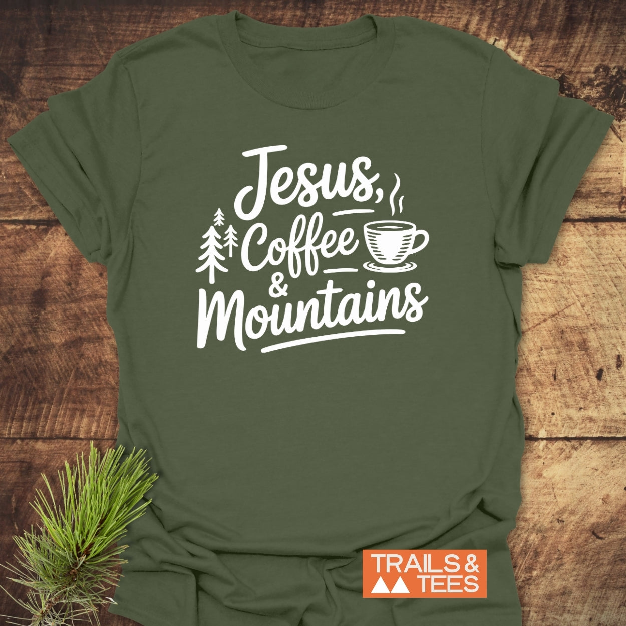 Jesus, Coffee, Mountains T-Shirt on rustic wood, featuring white text and pine tree and coffee cup drawings. Includes TRAILS & TEES logo and pine sprig, highlighting outdoor adventure spirit.