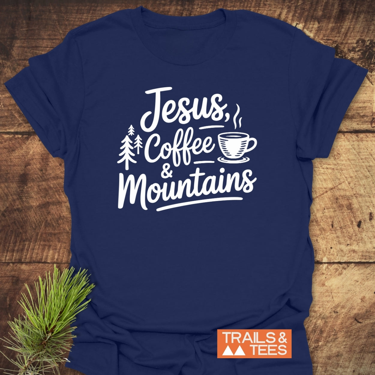 Jesus, Coffee, Mountains T-Shirt displayed on wood, featuring white script with a coffee cup and pine trees. Includes brand mark 'TRAILS & TEES' and styled with a pine sprig.