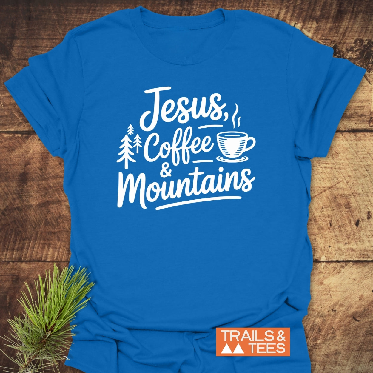 Jesus, Coffee, Mountains T-Shirt features a hand-lettered design with pine trees and a coffee cup graphic. Crafted from soft ring-spun cotton, it's styled on wood with a pine needle sprig.