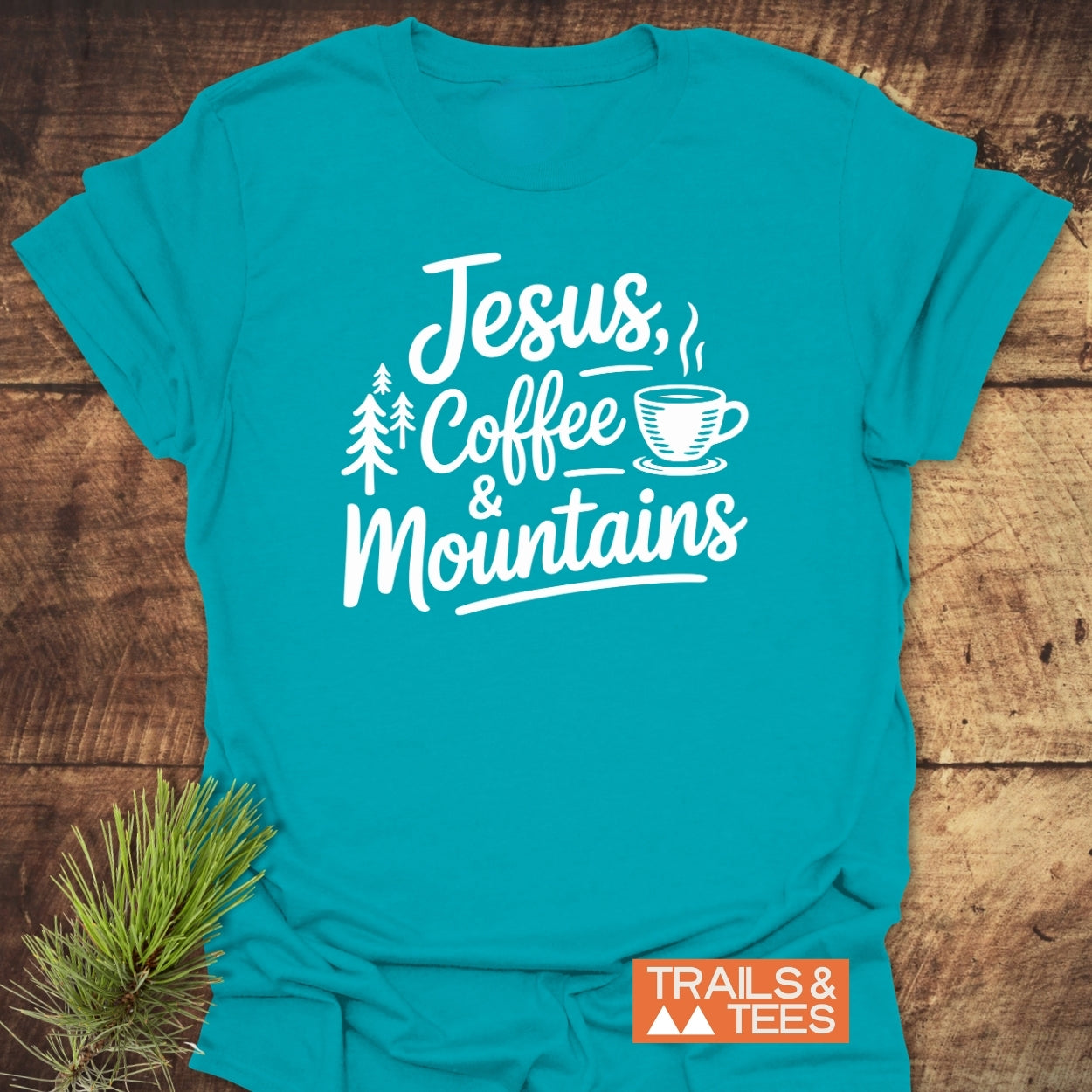 Jesus, Coffee, Mountains T-Shirt featuring white script and pine tree and coffee cup graphics on soft, ring-spun cotton. Includes a small TRAILS & TEES logo, laid on a wood surface.