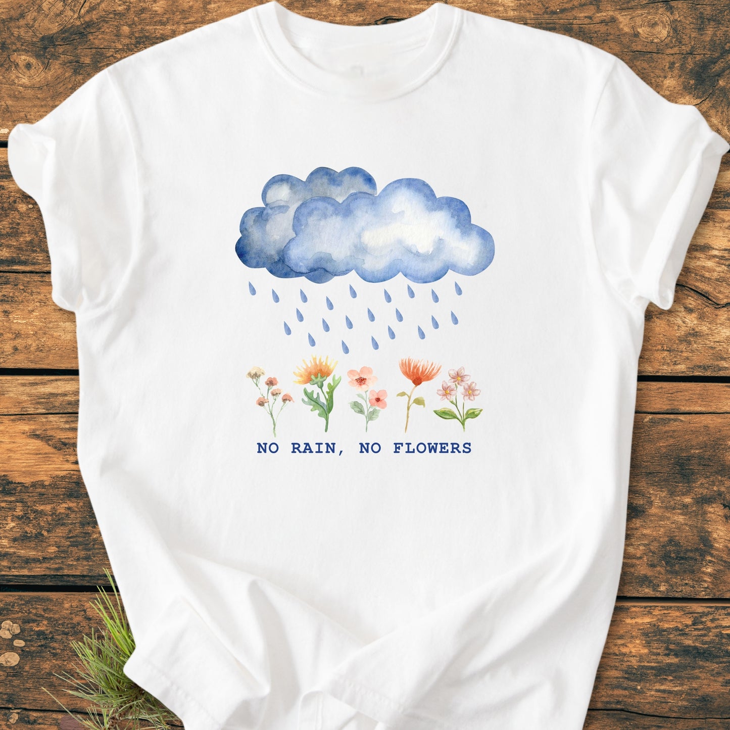 No Rain No Flowers - Vintage Heavyweight Tee features a watercolor-style blue rain cloud illustration above wildflowers with the slogan “NO RAIN, NO FLOWERS,” laid flat on a wooden surface.