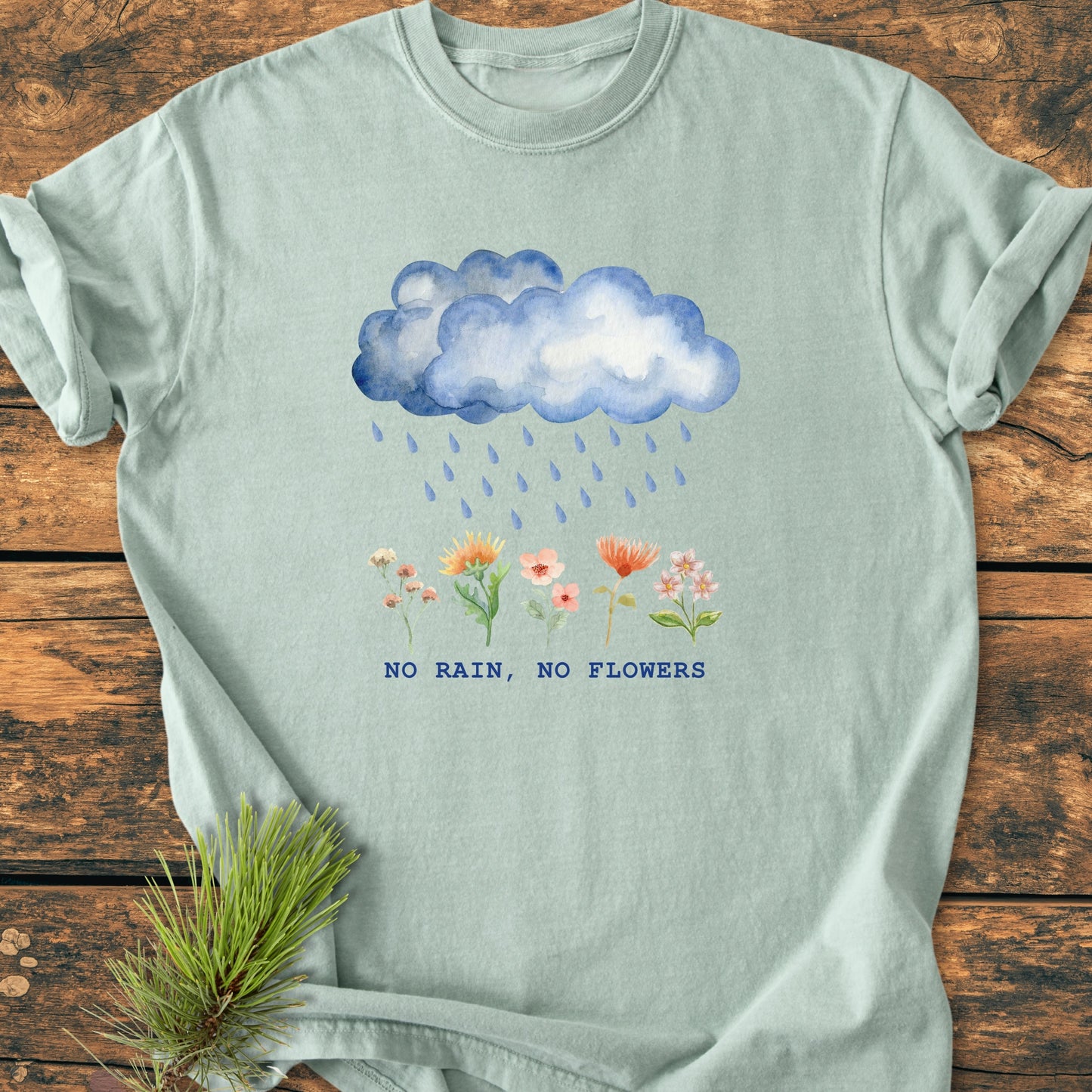 No Rain No Flowers - Vintage Heavyweight Tee features a watercolor blue rain cloud with flowers beneath, and bold lettering, laid flat on a wooden surface, highlighting its relaxed fit and durable stitching.