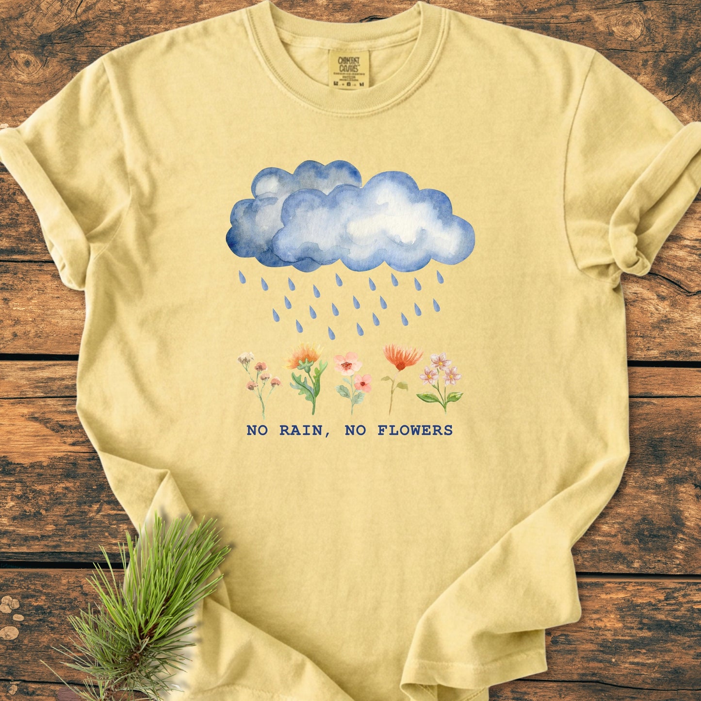 No Rain No Flowers - Vintage Heavyweight Tee features a watercolor-style graphic of raindrops on flowers with the slogan. Laid flat on a wooden surface, showcasing its relaxed fit and durable stitching.
