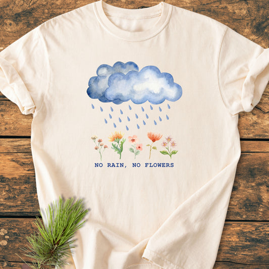 No Rain No Flowers - Vintage Heavyweight Tee features a watercolor rain cloud and flowers graphic with a slogan on a cream shirt, styled with pine needles on a wooden surface.