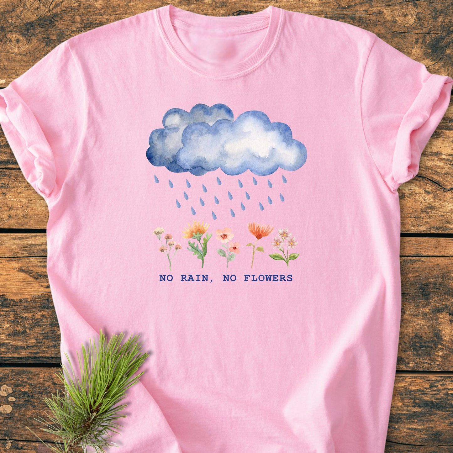 No Rain No Flowers - Vintage Heavyweight Tee: Soft pink T-shirt, watercolor blue cloud and raindrops, colorful flowers, NO RAIN, NO FLOWERS text, laid on wood with pine sprig beside.