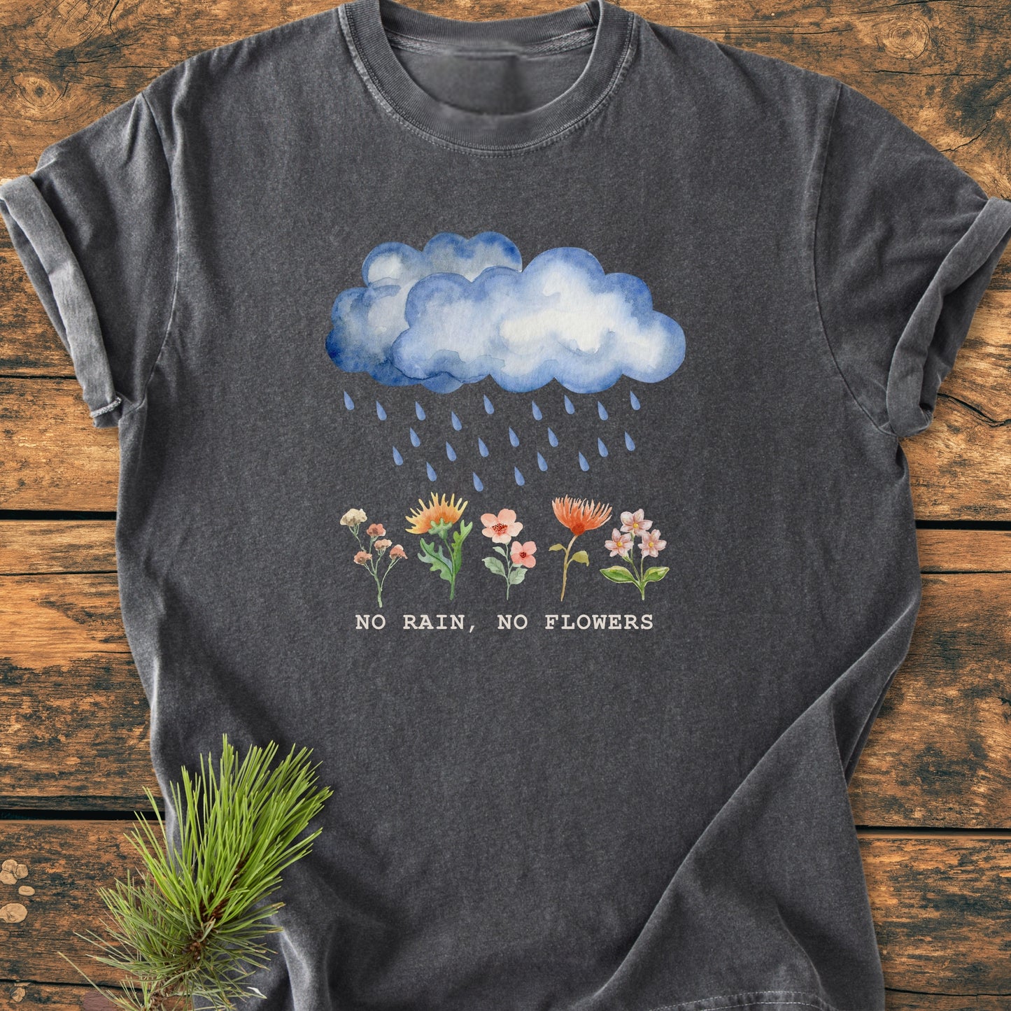 No Rain No Flowers - Vintage Heavyweight Tee features a watercolor cloud design above flowers, laid flat on wood. Crafted from soft, durable 100% ring-spun cotton, offering a relaxed fit.