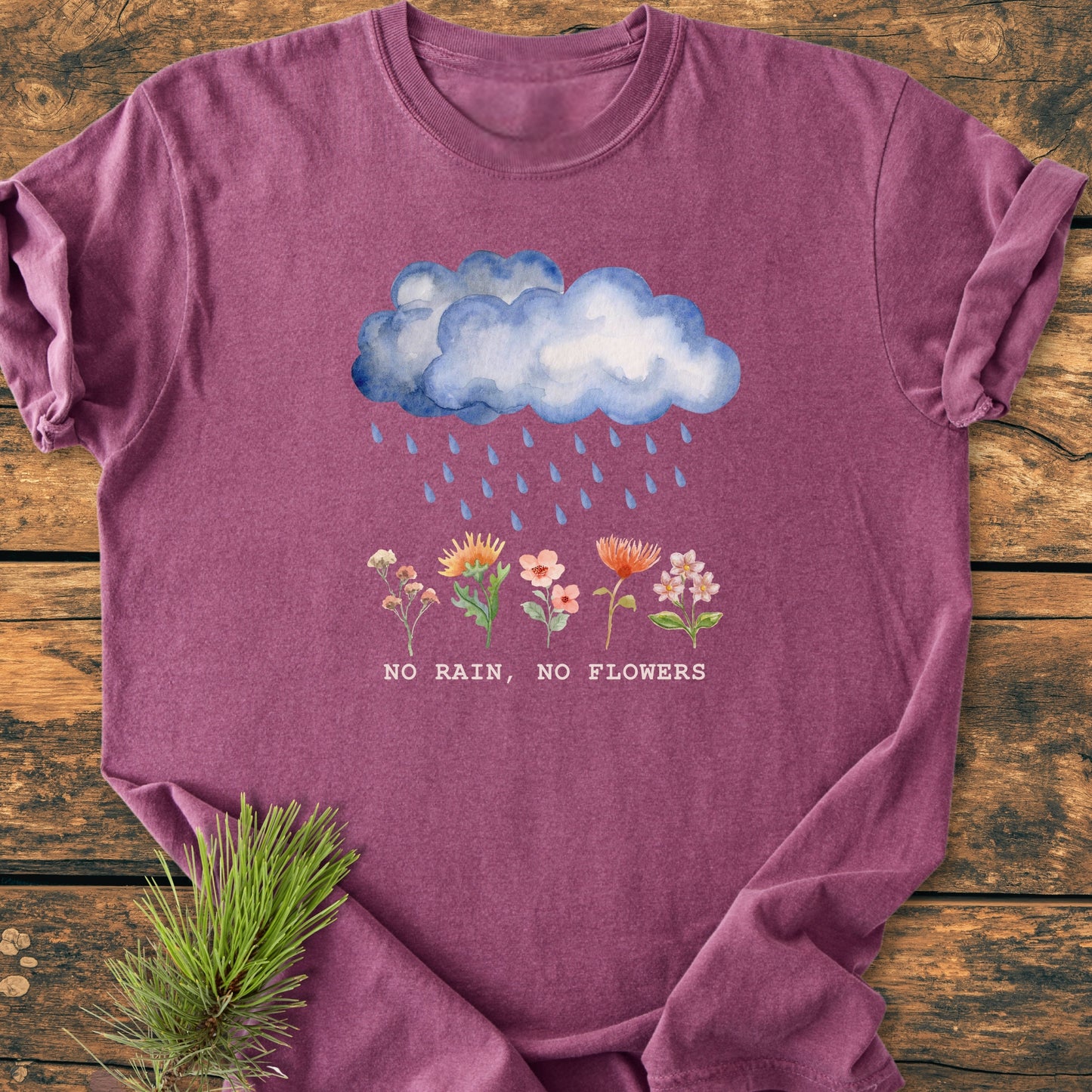 No Rain No Flowers - Vintage Heavyweight Tee features a watercolor rain cloud and flowers graphic with the slogan, laid on wood. Pine sprig accents the soft, durable, garment-dyed, relaxed-fit T-shirt.