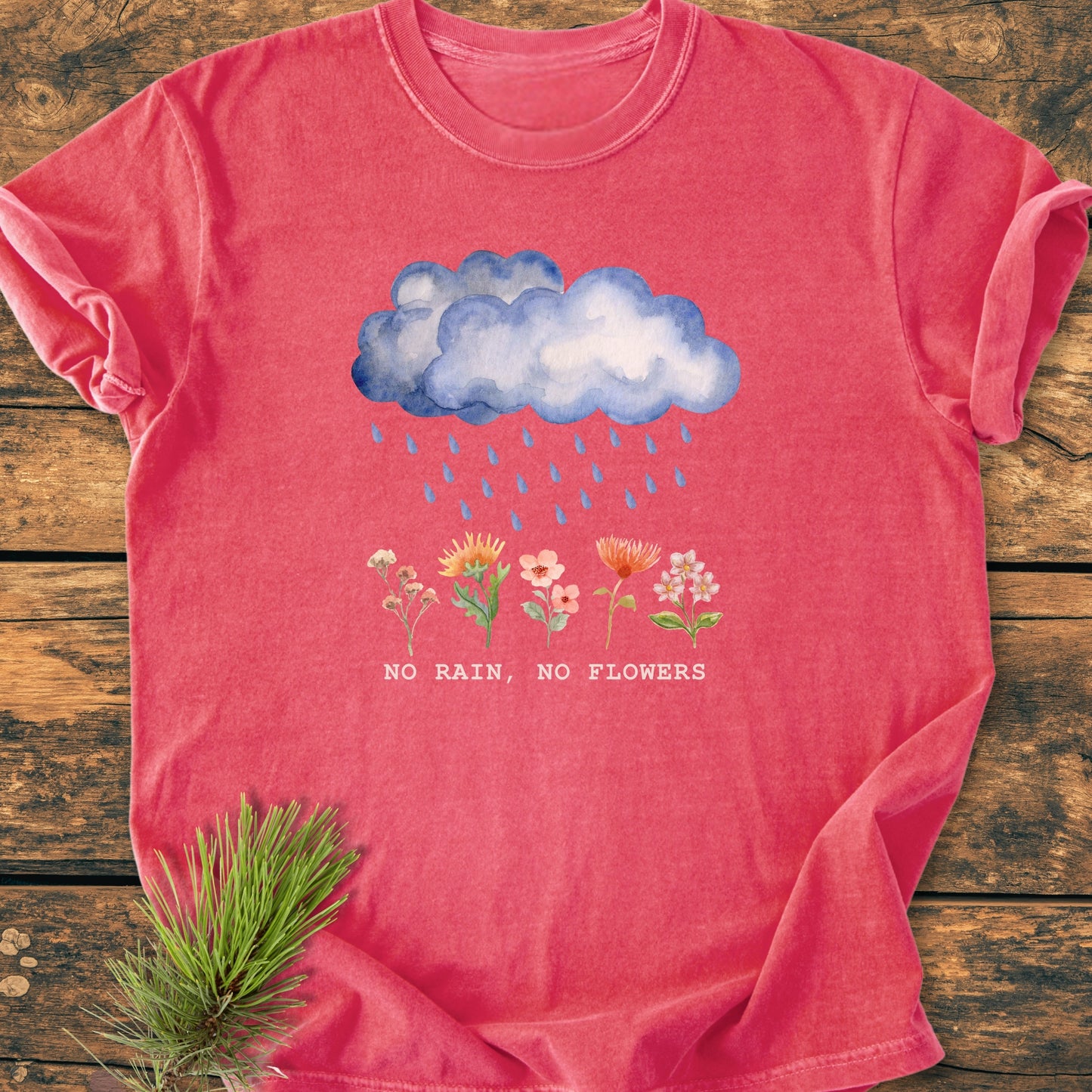 No Rain No Flowers - Vintage Heavyweight Tee featuring a watercolor rain cloud and blooming flowers illustration on a washed-out red background, laid on wood with pine needles beside it.