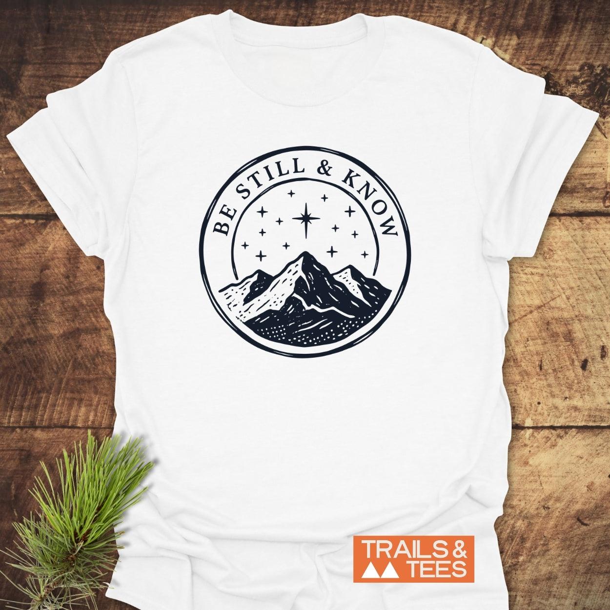 Be Still And Know T-Shirt featuring a mountain range under a starry sky, laid flat with pine accent; Trails & Tees logo in corner. Made of soft, lightweight cotton for outdoor enthusiasts.