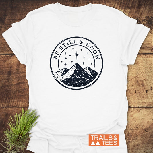 Be Still And Know T-Shirt featuring a mountain range under a starry sky, laid flat with pine accent; Trails & Tees logo in corner. Made of soft, lightweight cotton for outdoor enthusiasts.
