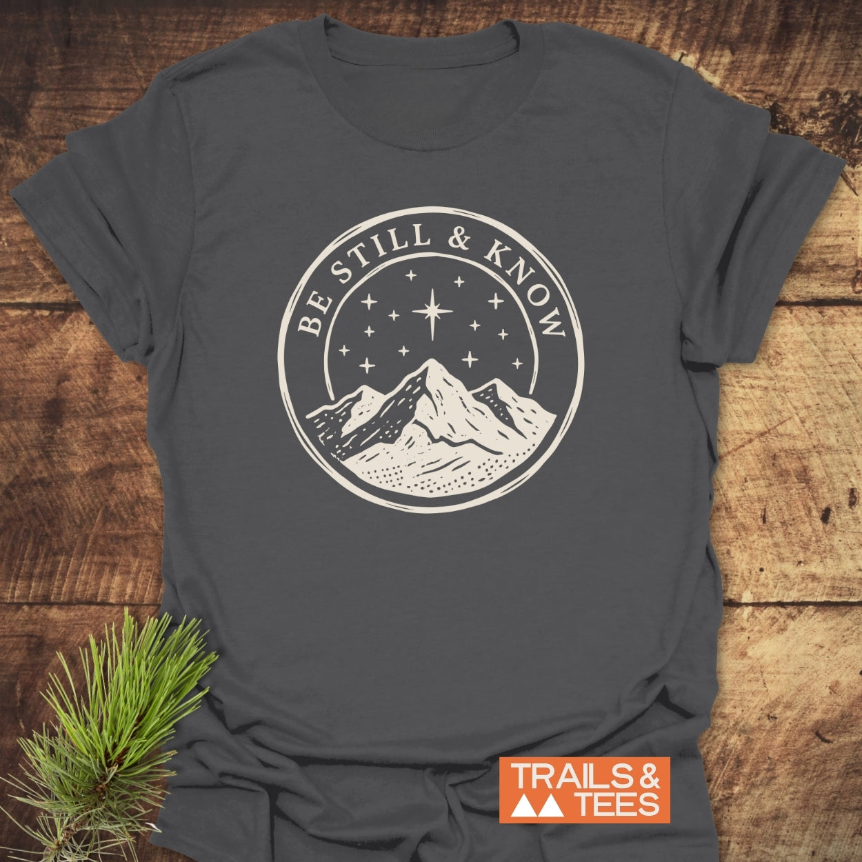 Charcoal-gray Be Still And Know T-Shirt featuring cream mountain graphic, laid on wood with pine sprig, exemplifies trail-tested comfort and timeless design, ideal for outdoor enthusiasts.