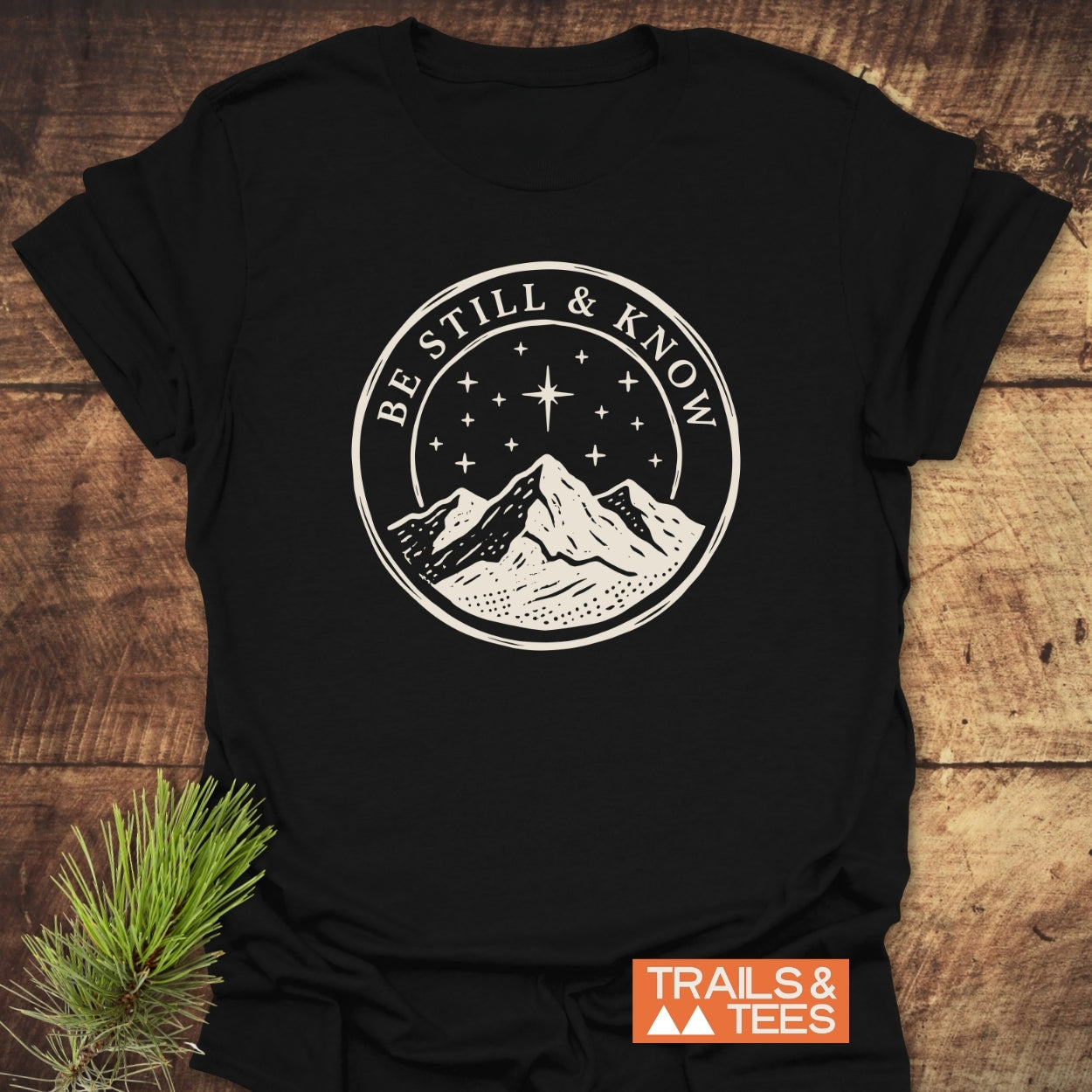 Be Still And Know T-Shirt featuring a cream mountain graphic under a starry sky, displayed on a wood surface with TRAILS & TEES logo, emphasizing comfort and adventure-ready design.