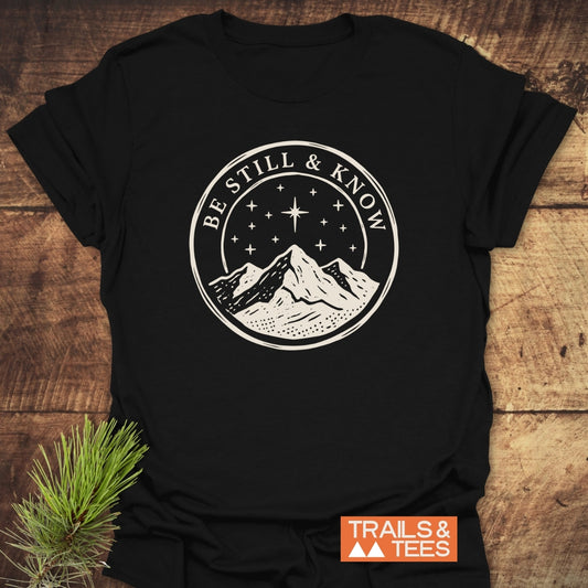 Be Still And Know T-Shirt featuring a cream mountain graphic under a starry sky, displayed on a wood surface with TRAILS & TEES logo, emphasizing comfort and adventure-ready design.