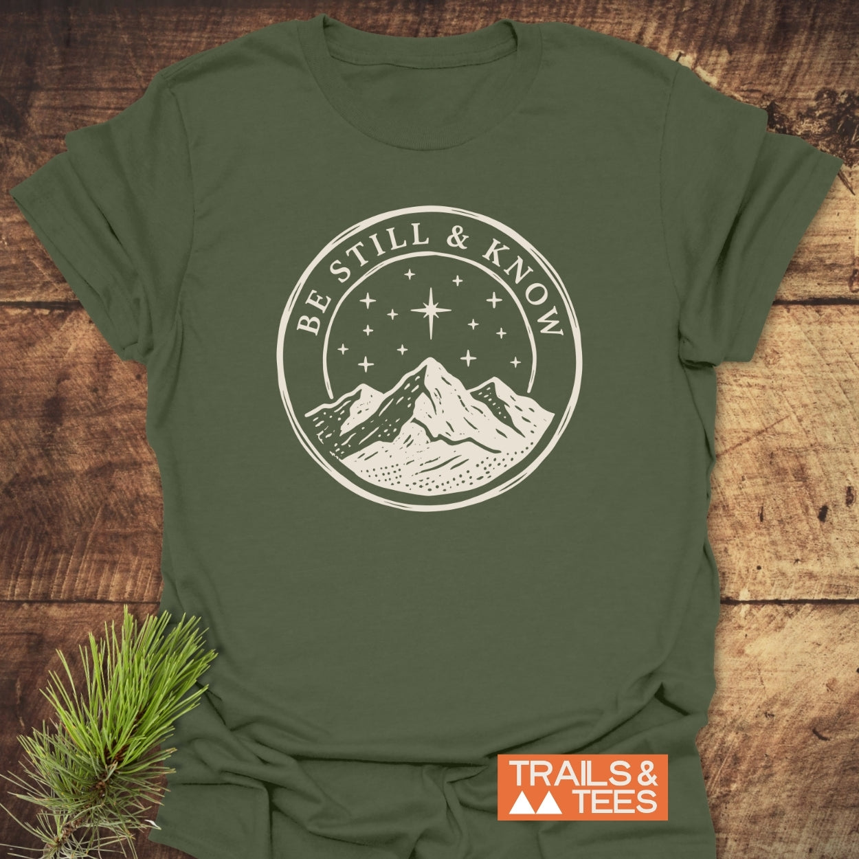 Be Still And Know T-Shirt featuring a mountain emblem under a starry sky, laid flat with a “TRAILS & TEES” label, accompanied by a pine sprig, showcasing outdoor-inspired design.