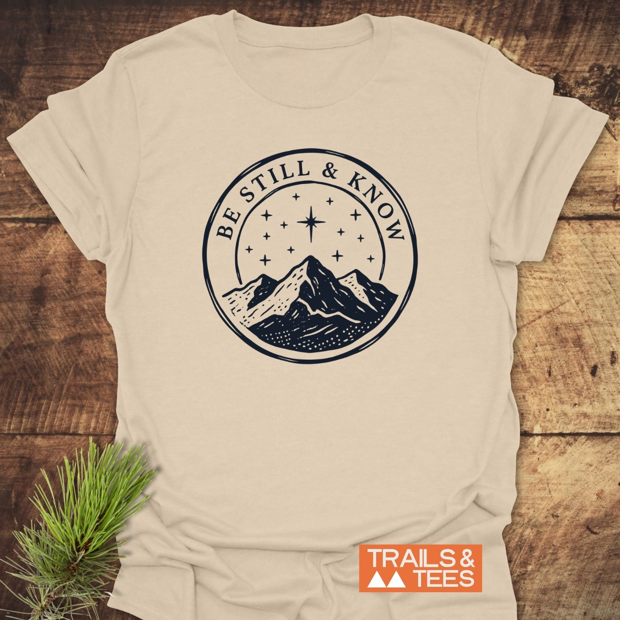 Light-beige Be Still And Know T-Shirt featuring a navy-blue emblem of a mountain under a starry sky, laid on wood with pine sprig, showcasing outdoor-inspired, comfortable design and quality craftsmanship.