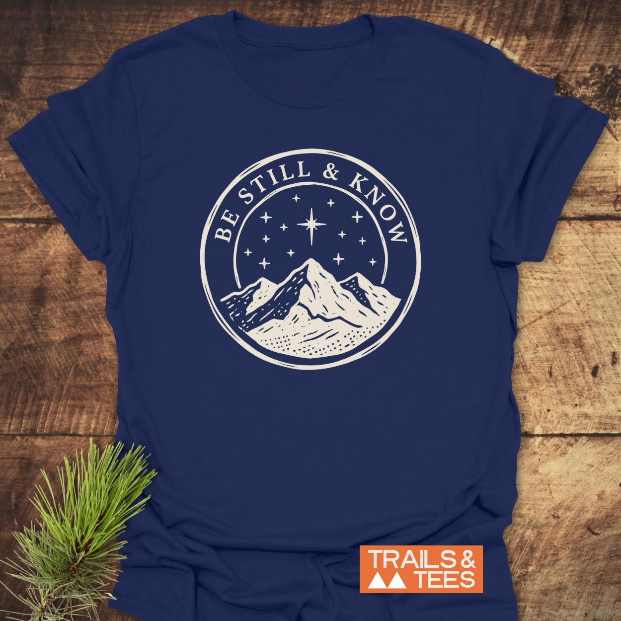 Be Still And Know T-Shirt featuring a white mountain graphic and starry sky on a navy-blue base, laid on wood. Includes the phrase BE STILL & KNOW inside the design's circle.