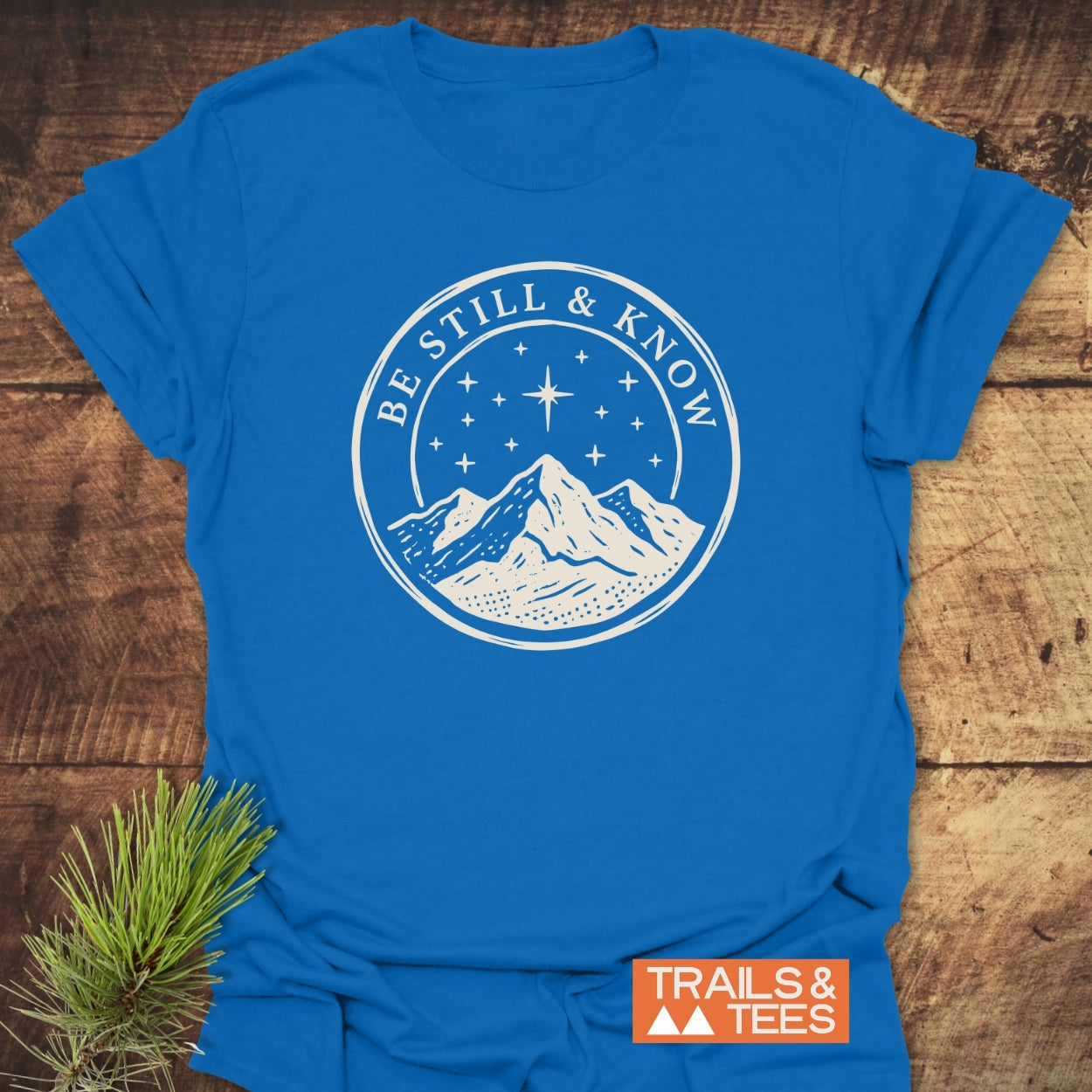 Be Still And Know T-Shirt with a mountain emblem under a starry sky, laid on wood with a pine sprig, embodying outdoor adventure and comfort in soft, ring-spun cotton.