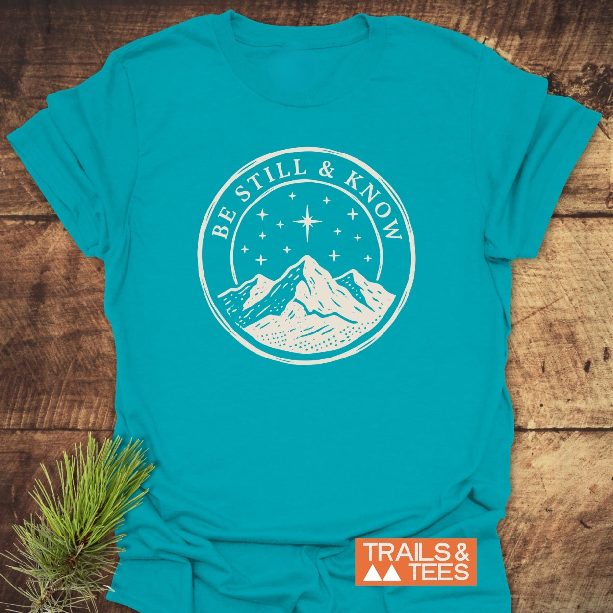 Be Still And Know T-Shirt featuring a mountain and starry sky graphic, laid flat with a pine sprig for decoration, showcasing comfort and adventure-ready design.