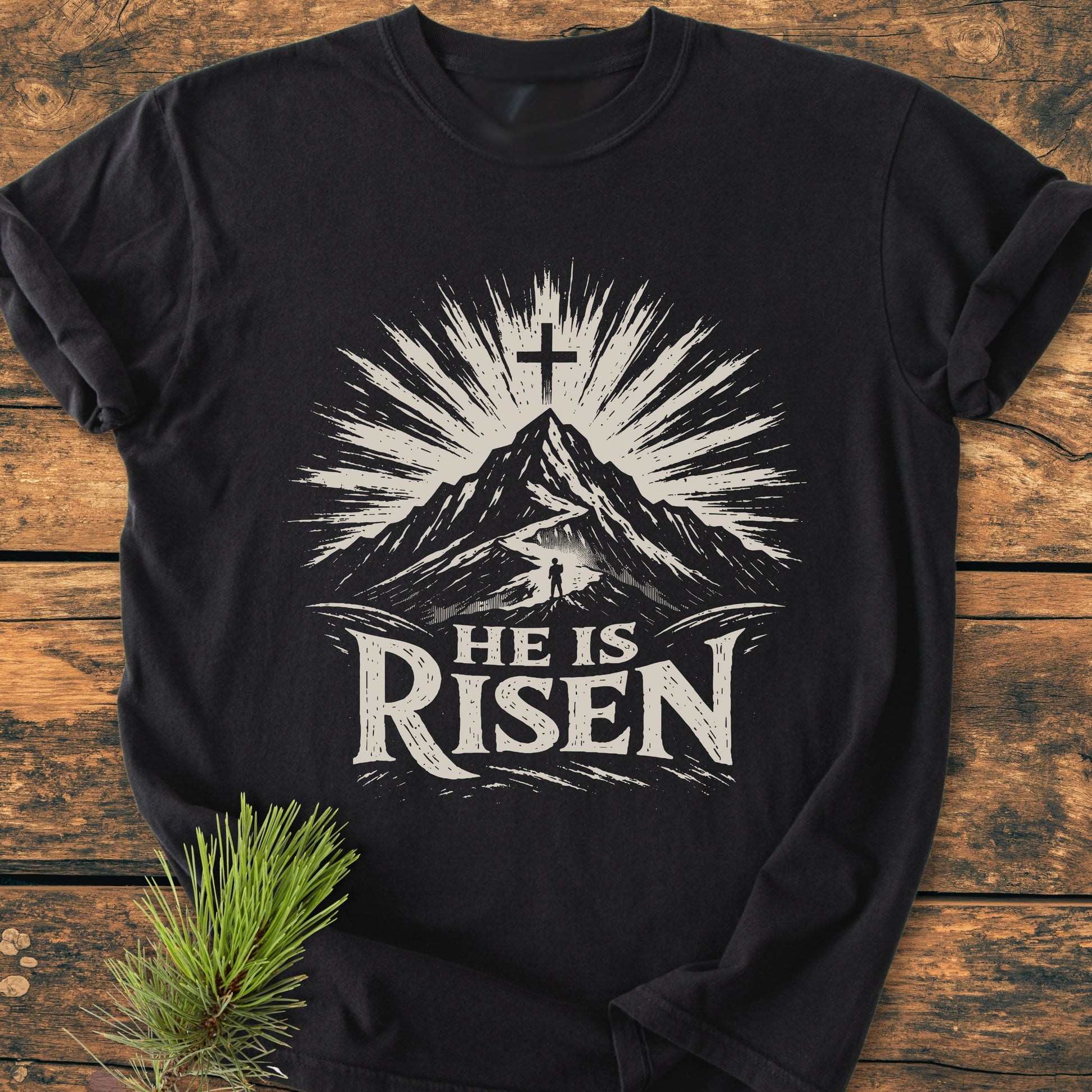 He Is Risen - Vintage Wash Tee features a mountain scene with a cross and sunburst, a lone figure, and rustic lettering on a black shirt, laid on wood beside a pine sprig.
