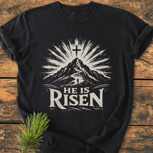 He Is Risen - Vintage Wash Tee features a mountain scene with a cross and sunburst, a lone figure, and rustic lettering on a black shirt, laid on wood beside a pine sprig.