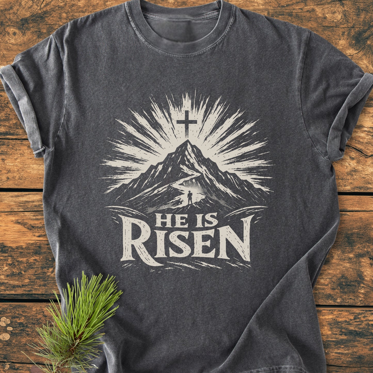He Is Risen - Vintage Wash Tee: Charcoal-gray T-shirt with a mountain and cross design, laid on a wooden surface, highlighting its relaxed fit and soft-washed texture for daily comfort.