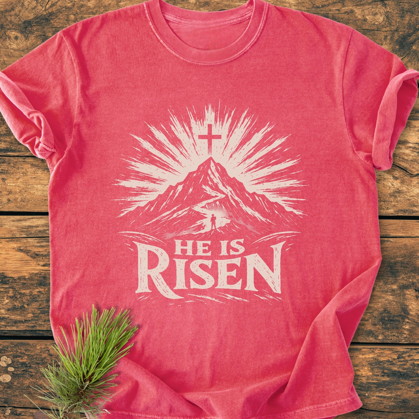 He Is Risen - Vintage Wash Tee features a mountain with a cross and rays, and a figure on a path, all in white graphics, on a soft, red garment-dyed T-shirt.