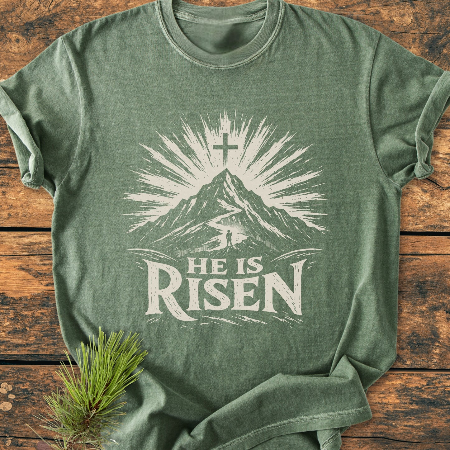 He Is Risen - Vintage Wash Tee displayed on wood, featuring a sunburst mountain graphic and text. Made of soft, durable 100% ring-spun cotton, offering comfort and a relaxed fit.