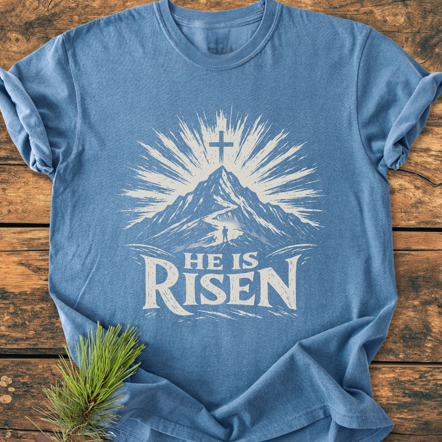 He Is Risen - Vintage Wash Tee features a stylized mountain with sunburst, path, and cross, plus bold lettering. Displayed flat on wood, complemented by a pine sprig.