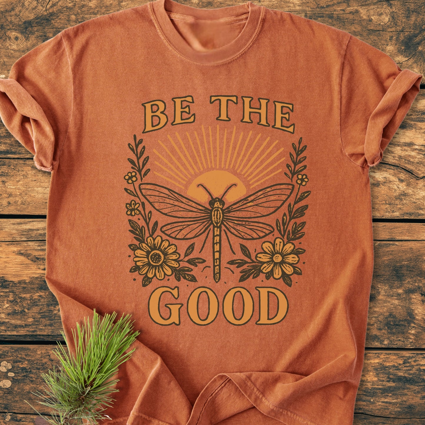 Be The Good - Vintage Wash Tee features a dragonfly graphic with a rising sun and flowers on a rust-orange, short-sleeved shirt, laid on a wooden surface with a pine sprig.