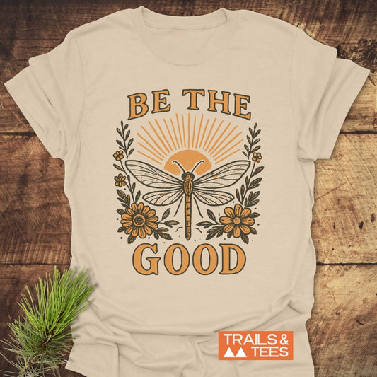 Be The Good T-Shirt featuring a dragonfly graphic with floral accents and BE THE GOOD text, laid flat on wood; includes TRAILS & TEES logo and pine branch detail.