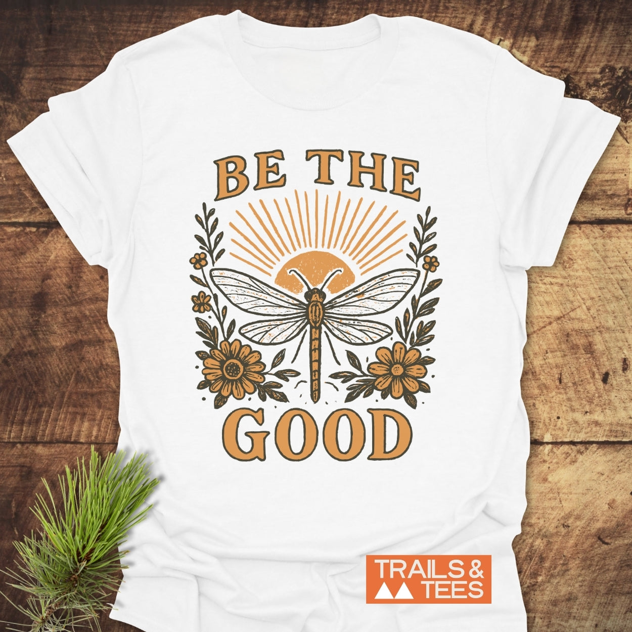 Be The Good T-Shirt features a dragonfly and sun design with floral accents, embodying outdoor spirit. Crafted from soft cotton, it showcases durability and comfort, perfect for everyday adventures.