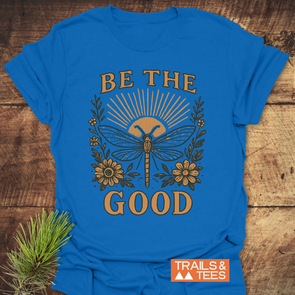 Be The Good T-Shirt featuring a dragonfly and rising-sun motif with floral accents, crafted from soft, lightweight cotton for trail-tested comfort. Displays TRAILS & TEES logo in the corner.