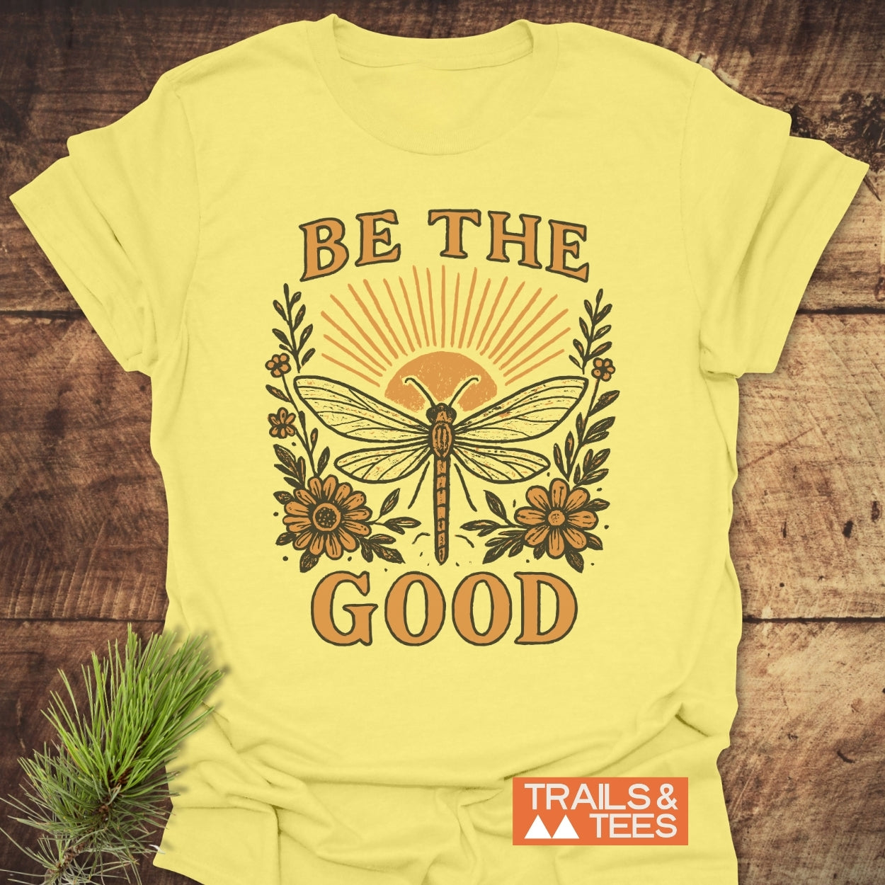 Be The Good T-Shirt featuring a dragonfly and sun design with floral accents, showcasing BE THE GOOD. Made from soft, lightweight cotton, perfect for outdoor adventures or casual wear. Logo: TRAILS & TEES.