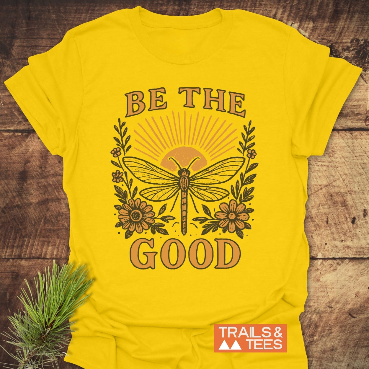 Be The Good T-Shirt with dragonfly and floral design, featuring a rising-sun motif. Made from soft, ring-spun cotton for comfort, includes TRAILS & TEES logo near hem.