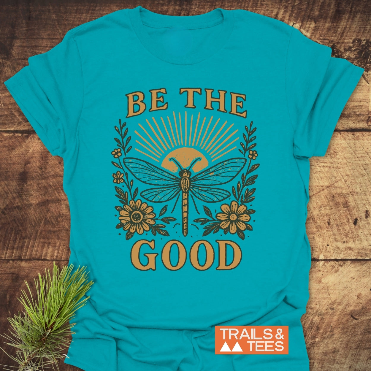 Be The Good T-Shirt with a dragonfly and rising sun graphic, surrounded by flowers. Crafted from soft cotton for adventure, featuring durable prints and a logo. Pine sprig accents the display.