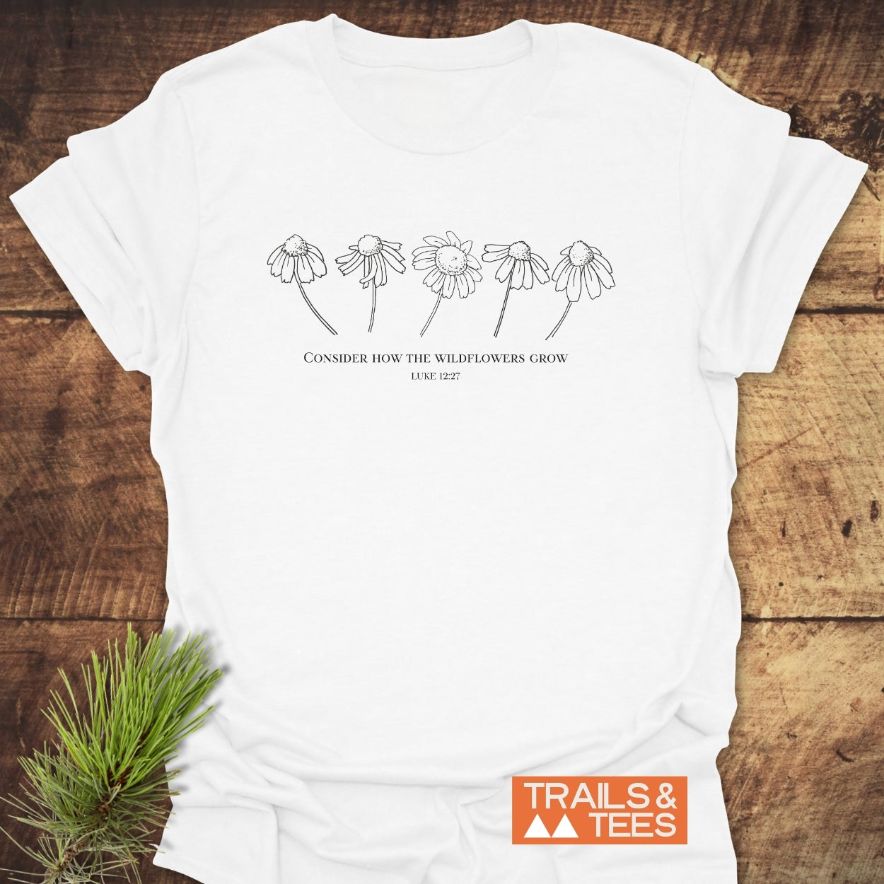 Consider How The Wildflowers Grow T-Shirt on rustic wood, featuring five daisylike wildflowers with the quote and citation. Orange TRAILS & TEES logo in corner, emphasizing outdoor-inspired comfort and design.