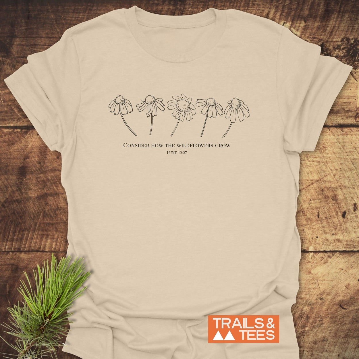 Consider How The Wildflowers Grow T-Shirt featuring a line drawing of wildflowers and a quote from Luke 12:27, styled with a sprig of evergreen on a wooden surface.