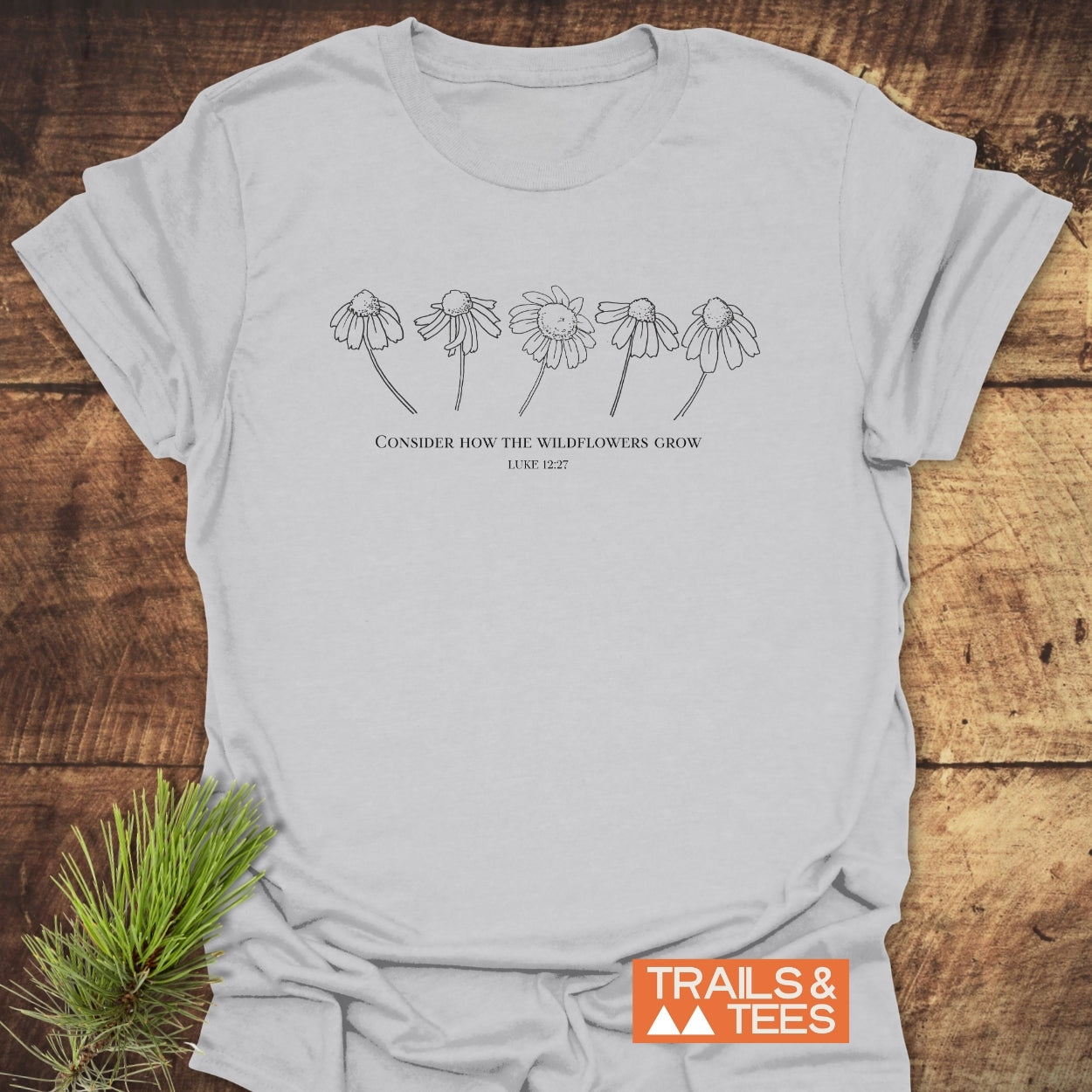 Consider How The Wildflowers Grow T-Shirt featuring five daisy-like wildflowers and biblical quote, styled with a pine sprig on a wooden surface, emphasizing comfort and durability by Trails & Tees.