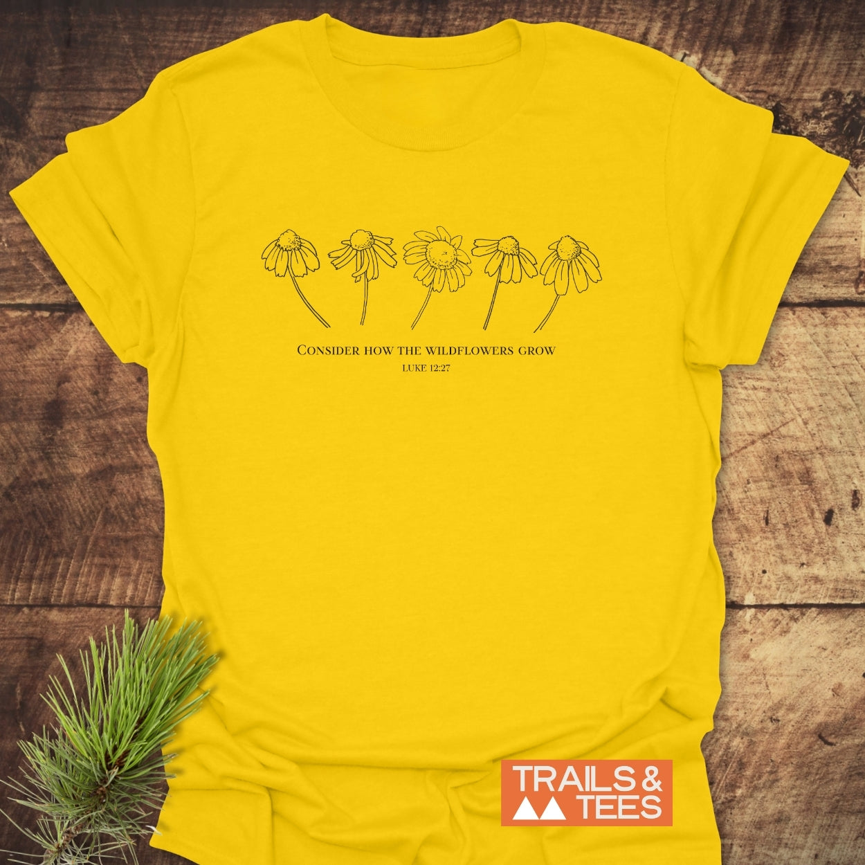 Consider How The Wildflowers Grow T-Shirt featuring black wildflower line art and scripture on soft yellow cotton, laid on wood with pine branch, Trails & Tees logo visible.