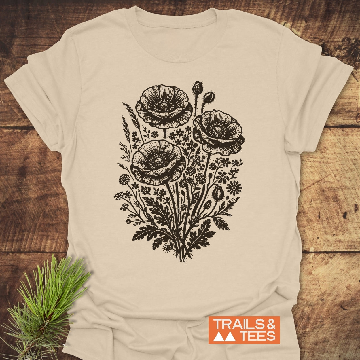 Wildflower Bouquet T-Shirt featuring a hand-drawn botanical print with poppy blooms and wildflowers. Includes TRAILS & TEES logo. Laid on wood, highlighting its soft, lightweight cotton construction.