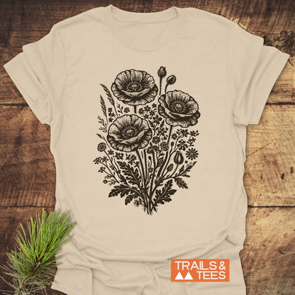 Wildflower Bouquet T-Shirt featuring a hand-drawn botanical print with poppy blooms and wildflowers. Includes TRAILS & TEES logo. Laid on wood, highlighting its soft, lightweight cotton construction.