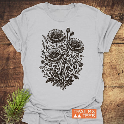 Wildflower Bouquet T-Shirt on wooden surface, showcasing a black botanical illustration of poppies and wildflowers. TRAILS & TEES logo visible, emphasizing outdoor-inspired comfort and timeless design.