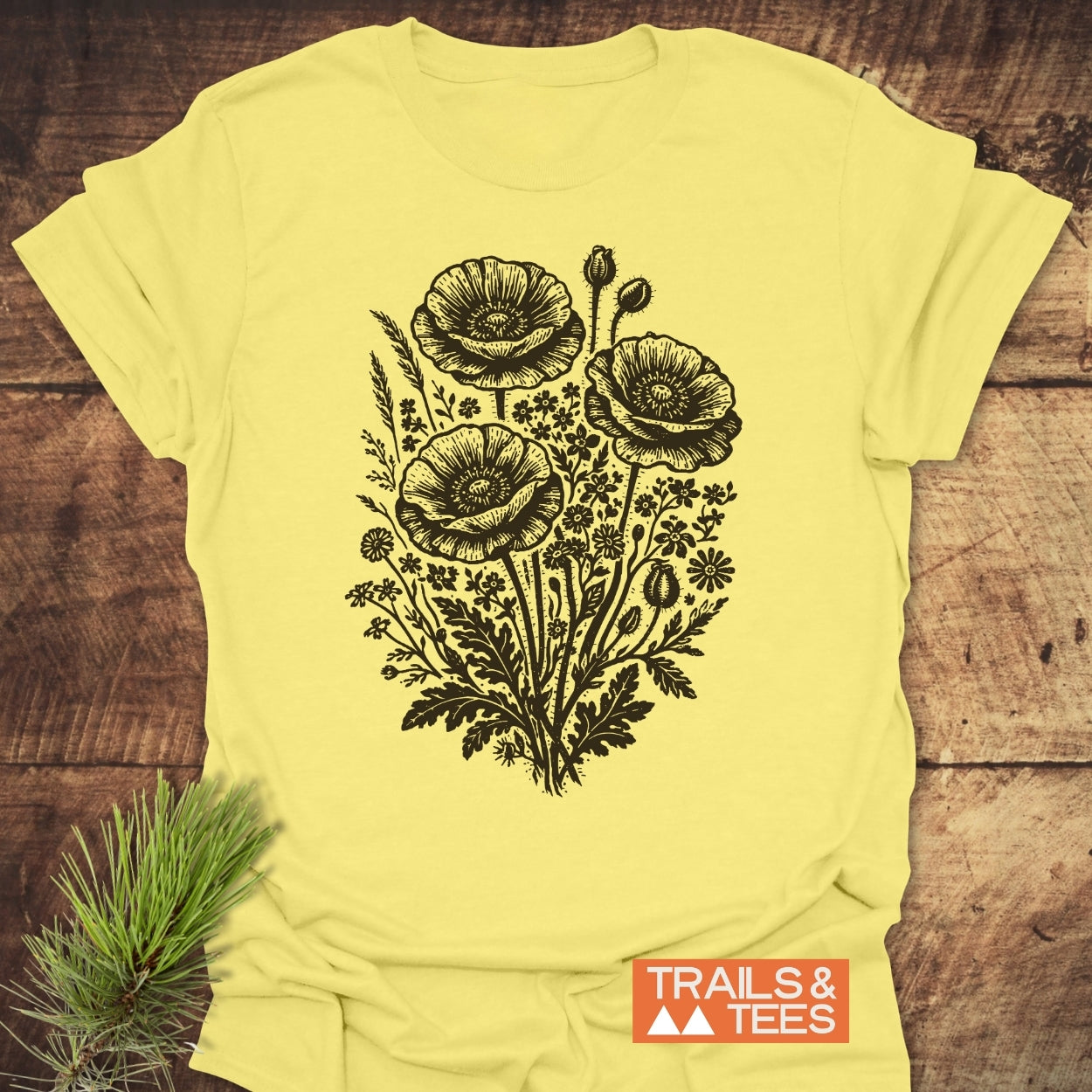 Wildflower Bouquet T-Shirt features a black line-art bouquet of poppy-like blooms on a pale yellow background, with a TRAILS & TEES logo, laid on rustic wood.