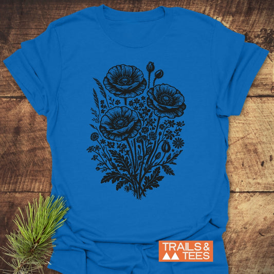 Wildflower Bouquet T-Shirt featuring a detailed black botanical illustration of poppies on a bright blue fabric, laid on rustic wood with a pine sprig beside it and orange TRAILS & TEES logo visible.