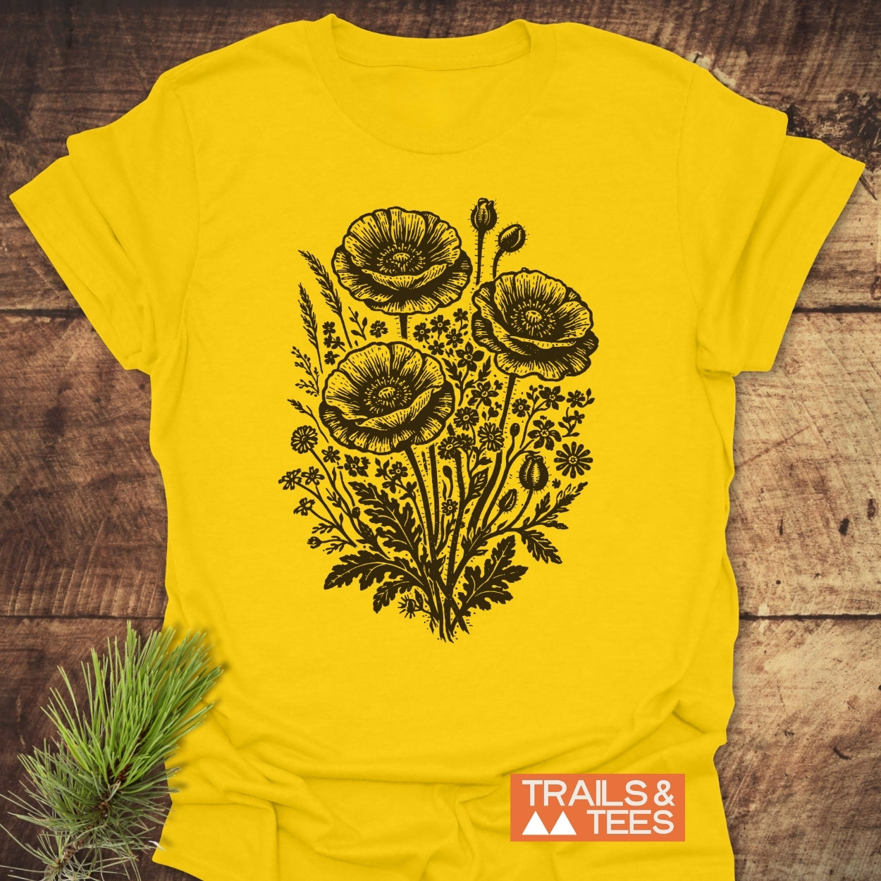 Wildflower Bouquet T-Shirt featuring a black botanical illustration of poppy blossoms. Laid flat on wood with a pine sprig. Logo TRAILS & TEES visible, highlighting outdoor-inspired design.
