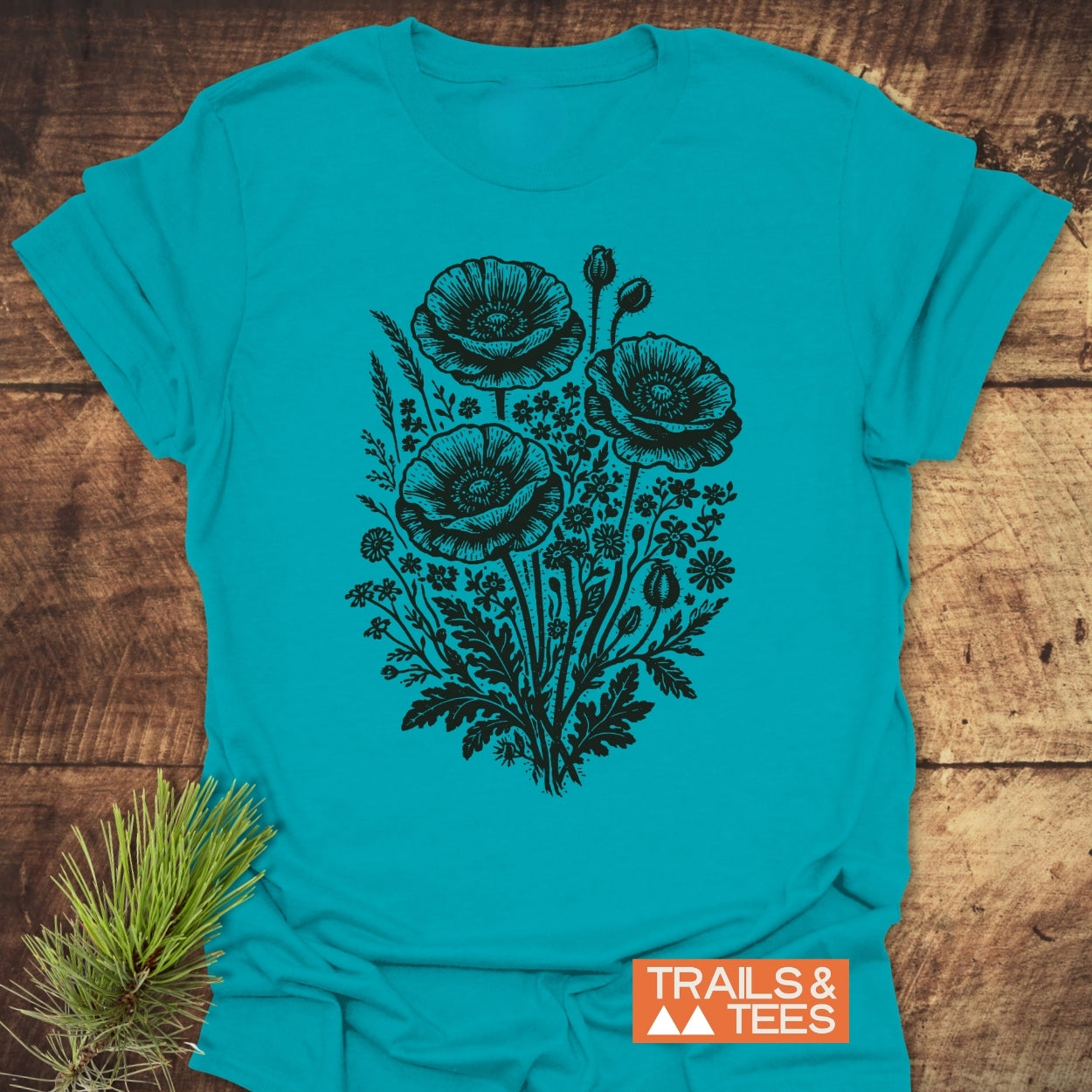 Wildflower Bouquet T-Shirt showcasing a stylized wildflower design with poppy-style blooms, laid flat beside a spruce twig on wood. Features the TRAILS & TEES logo, highlighting its outdoor-inspired appeal.