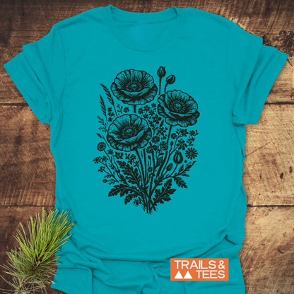 Wildflower Bouquet T-Shirt showcasing a stylized wildflower design with poppy-style blooms, laid flat beside a spruce twig on wood. Features the TRAILS & TEES logo, highlighting its outdoor-inspired appeal.