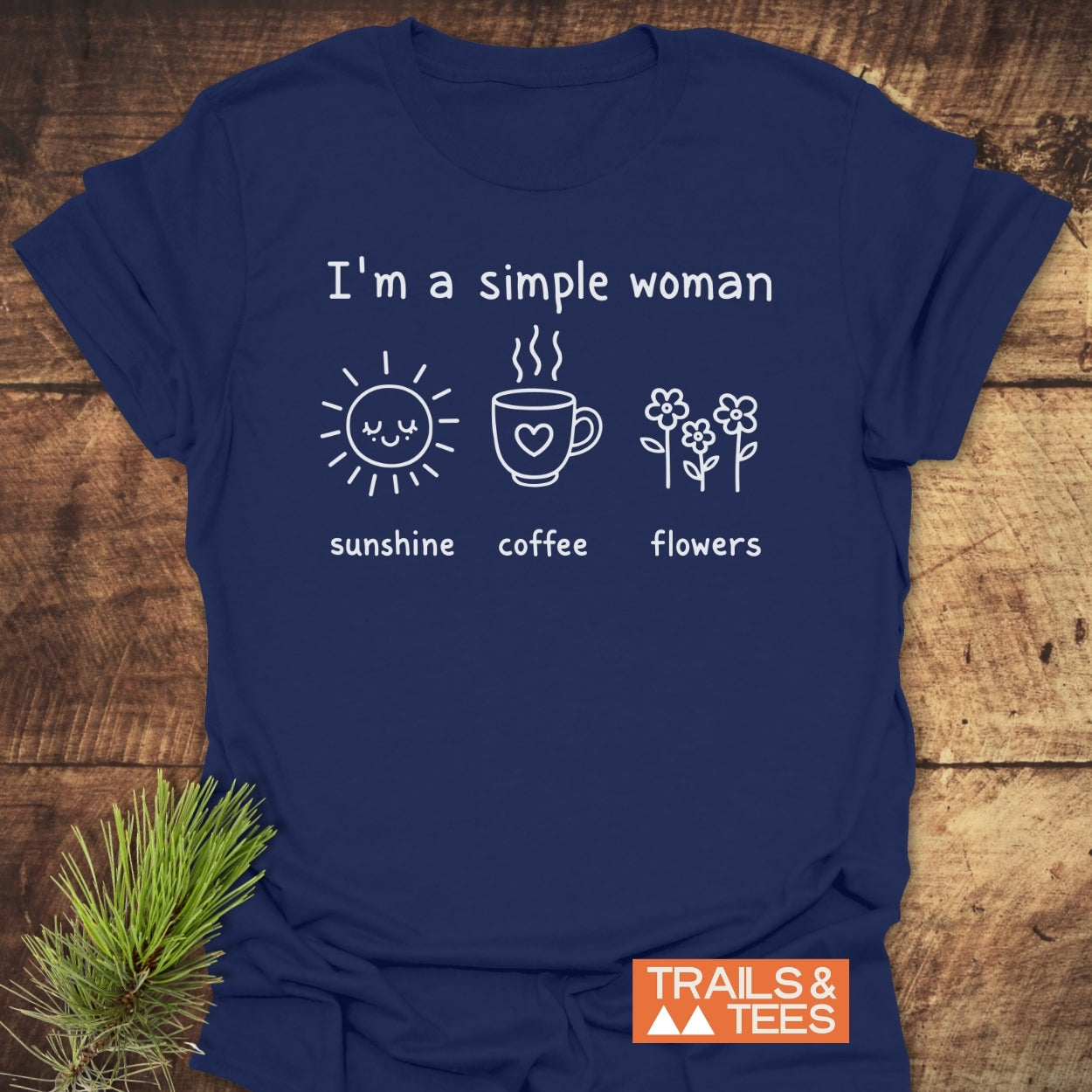 I'm A Simple Woman. Sunshine Coffee Flowers T-Shirt on wooden surface, featuring white print with sun, coffee mug, flowers icons, and TRAILS & TEES logo. Pine sprigs accent the scene.